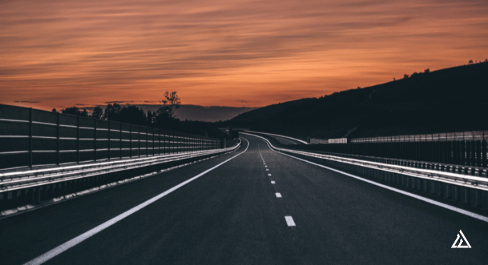 Image of a highway paved with asphalt. The asphalt is new, since it is very black. The road keeps going into the distance against a pink and orange sunset.