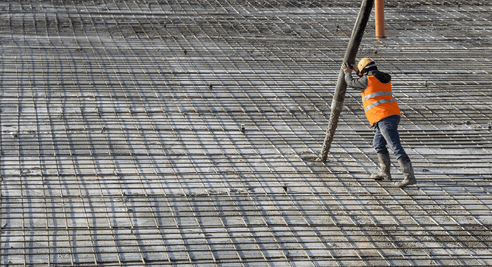 The image displays a construction worker using a concrete pump to place fresh concrete on a foundation. 