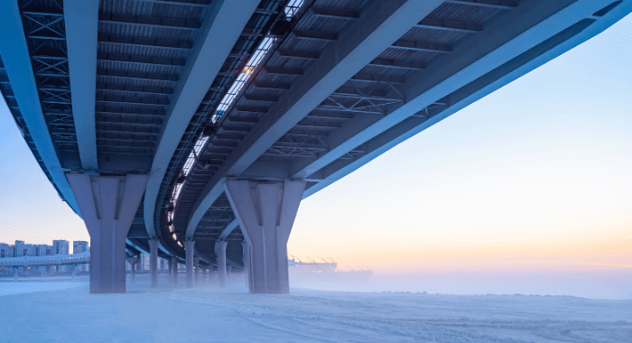 The image shows the underside of a bridge. The angle displays the concrete support of the structure amid snow. For this concrete structure to survive harsh weathers, an air-entraining agent should be added to the concrete mix design. This decision would increase concrete's resistance to freeze thaw.